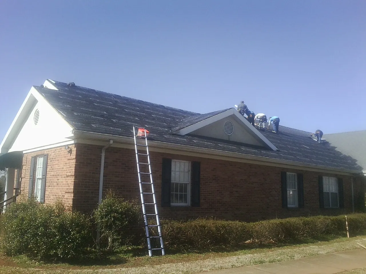 Crew tearing shingles off a brick ranch roof with a ladder set — Emergency Roof Repair work by our crew in Ladson
