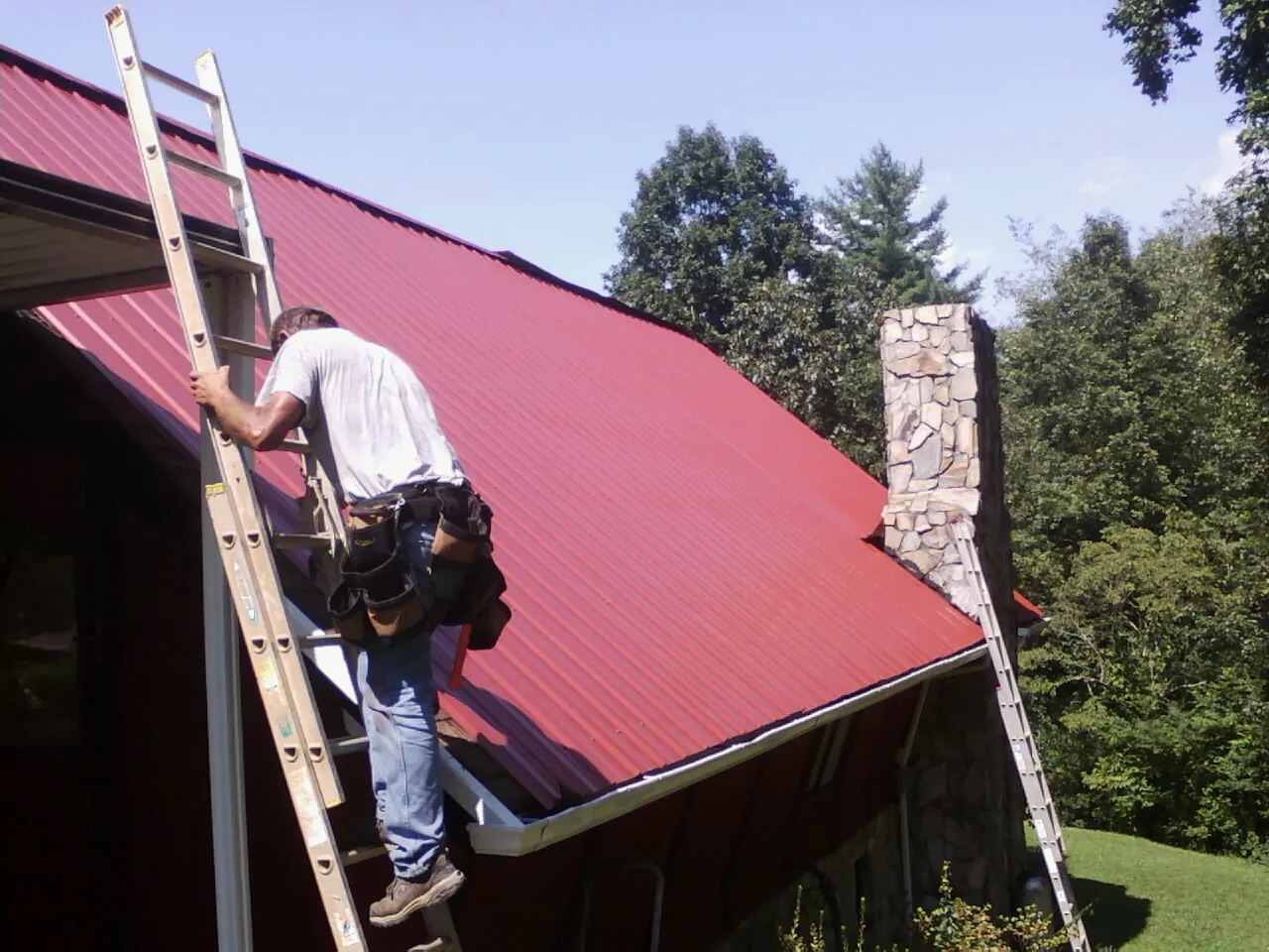Roofer on a ladder beside a red metal roof and stone chimney — Soffit & Fascia Repair by Roofing Brothers in Ladson